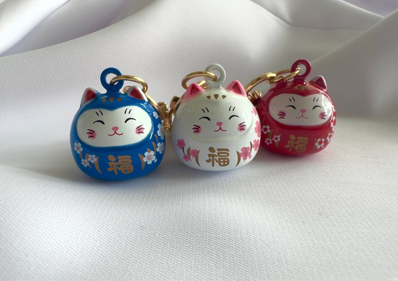 May include: Three lucky cat charms with gold accents. The charms are blue, white, and red with pink cherry blossoms and the word "福" (meaning "good fortune") in gold.
