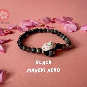 Könnte beinhalten: Schwarzes Perlenarmband mit einem weißen Maneki-Neko-Anhänger. Der Anhänger hat ein schwarzes und rotes Design und ist vor einem rosa Hintergrund platziert.