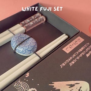 Op de afbeelding: Een set witte eetstokjes met een blauwe en witte keramische eetstokjeshouder in een zwarte doos. De doos heeft Japanse tekst die "食卓を彩る。" luidt, wat vertaald wordt als "Decoreer de tafel." De doos heeft ook de tekst "WeebsLab" erop gedrukt.