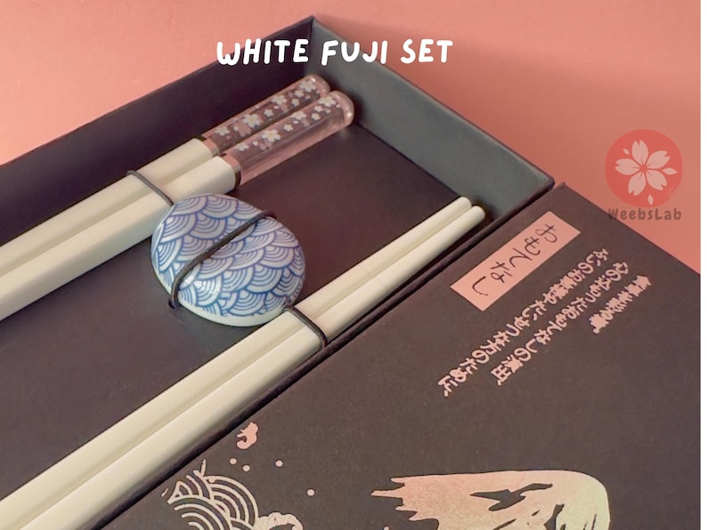 Op de afbeelding: Een set witte eetstokjes met een blauwe en witte keramische eetstokjeshouder in een zwarte doos. De doos heeft Japanse tekst die "食卓を彩る。" luidt, wat vertaald wordt als "Decoreer de tafel." De doos heeft ook de tekst "WeebsLab" erop gedrukt.