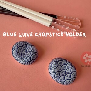 Op de afbeelding: Twee blauwe en witte keramische eetstokjeshouders met een golfpatroon. De houders staan op een roze oppervlak. De tekst "BLUE WAVE CHOPSTICK HOLDER" is zichtbaar in de afbeelding.