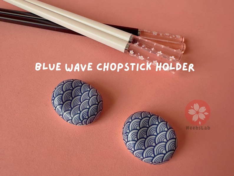Op de afbeelding: Twee blauwe en witte keramische eetstokjeshouders met een golfpatroon. De houders staan op een roze oppervlak. De tekst "BLUE WAVE CHOPSTICK HOLDER" is zichtbaar in de afbeelding.