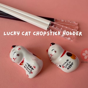 Op de afbeelding: Twee witte keramische gelukkatten-eetstokjeshouders met rode en gouden accenten. De katten liggen met opgeheven poten. De tekst op de houders luidt "Veel geluk, veel geluk, veel geluk".