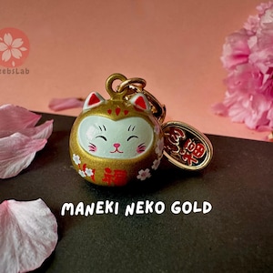May include: A gold Maneki Neko charm with white and red details. The charm features a smiling cat face and the Japanese word "福" (meaning "good fortune") on the front. The charm is attached to a gold chain with a small gold tag that says "招福" (meaning "invite good fortune").