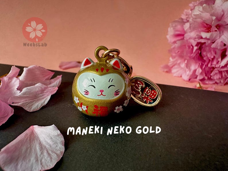 May include: A gold Maneki Neko charm with white and red details. The charm features a smiling cat face and the Japanese word "福" (meaning "good fortune") on the front. The charm is attached to a gold chain with a small gold tag that says "招福" (meaning "invite good fortune").