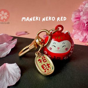 May include: A red Maneki Neko, or lucky cat, keychain with a gold charm that says "福" (meaning "good fortune") in Japanese. The keychain is surrounded by pink cherry blossom petals.