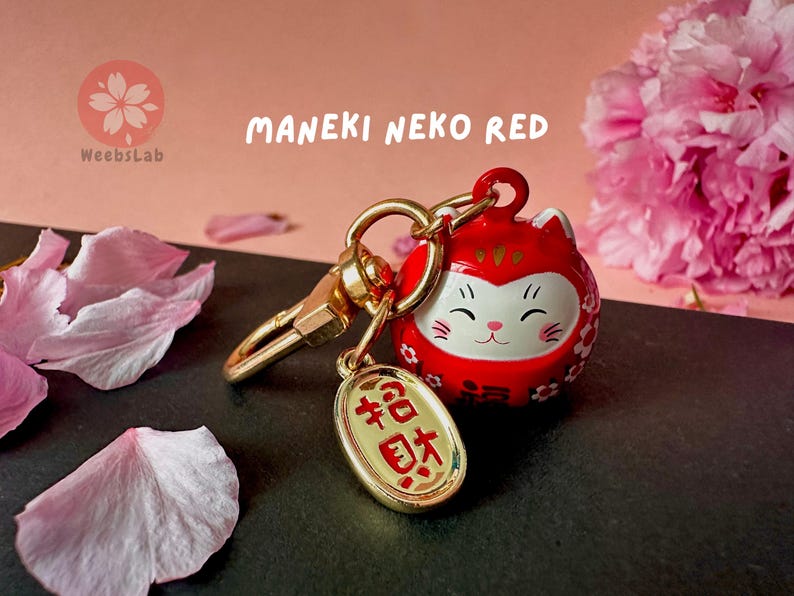 May include: A red Maneki Neko, or lucky cat, keychain with a gold charm that says "福" (meaning "good fortune") in Japanese. The keychain is surrounded by pink cherry blossom petals.
