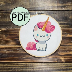 Puede incluir: Patrón de punto de cruz PDF para un gato blanco con un cuerno de unicornio rosa y dorado. El gato está sentado con la cola enroscada detrás de él.