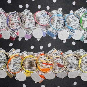 Può includere: Una collezione di venti portachiavi con anelli in silicone colorati e anelli in metallo argentato. Ogni portachiavi ha un'etichetta bianca con un numero. I portachiavi sono disposti su una superficie nera a pois bianchi.