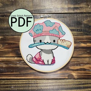May include: Cross stitch pattern PDF of a cute cartoon cat wearing a pink and blue shower cap and holding a toothbrush. The cat is on a white background.