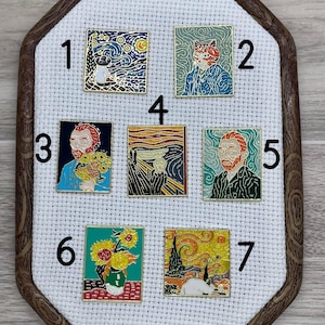 May include: Seven enamel pins featuring famous paintings by Vincent van Gogh. The pins are displayed on a white fabric background with numbers 1 through 7. The pins include a cat in a starry night, a cat in a swirling blue background, a self-portrait of van Gogh, a painting of a cypress tree, a self-portrait of van Gogh, sunflowers in a vase, and a starry night with a cat.
