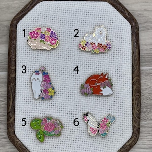 May include: Six enamel pins featuring various animals and floral designs. The pins are displayed on a white fabric background with numbers 1 through 6 next to each pin.