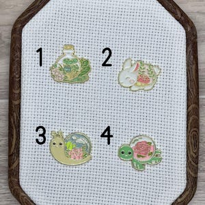 May include: Four enamel pins featuring a glass bottle with a plant inside, a rabbit with a flower, a snail with a flower, and a turtle with a flower. The pins are numbered 1 through 4.
