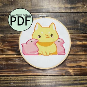 Op de afbeelding: Een kruissteekpatroon in een houten hoepel toont een gele kat met een gele kraag en twee roze marshmallow kuikens. De tekst "Cross Stitch Pattern PDF" staat in een groene cirkel linksboven.