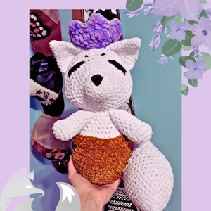 May include: A handmade crochet arctic fox plush toy in a flower pot. The fox is white with black details and a purple crown. The flower pot is brown. The text "Arctic Fox in a Flower Pot" is at the top of the image.