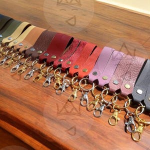 Könnte beinhalten: Eine Sammlung von Leder-Schlüsselanhängern in verschiedenen Farben, darunter Orange, Grau, Grün, Braun, Rot und Lila. Jeder Schlüsselanhänger hat einen Metallverschluss und einen Metallring. Die Schlüsselanhänger sind auf einer Holzoberfläche angeordnet.