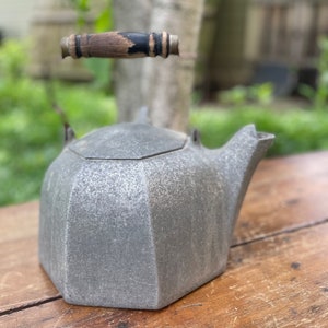 May include: A gray cast iron teapot with a wooden handle. The teapot has a geometric shape and a textured surface.
