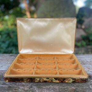 May include: An open vintage jewelry box with a gold trim and a marbled pattern on the bottom. The box has 16 compartments lined with a soft brown fabric.