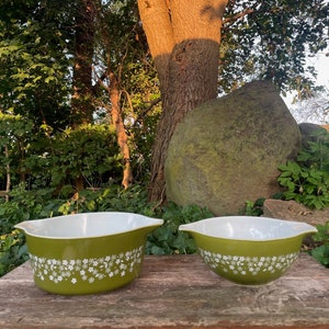 May include: Two green mixing bowls with white floral patterns on a wooden surface. The bowls are in a natural outdoor setting with a large tree and rock in the background.