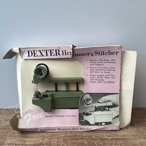 Może przedstawiać: Zielona maszyna do szycia Dexter Automatic Hemmer and Stitcher w oryginalnym pudełku. Pudełko jest białe z różowym i czarnym tekstem. Maszyna jest pokazana w użyciu, podczas obszywania kawałka materiału. Pudełko wymienia takie funkcje, jak obszywanie, szycie zygzakiem, szycie zygzakiem, przyszywanie guzików i zamków błyskawicznych, szycie niewidoczne i obszywanie zasłon i firan.