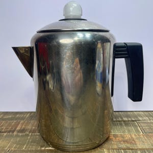 Puede incluir: Una cafetera de percolación de plata con un asa negra y una perilla blanca en la tapa.