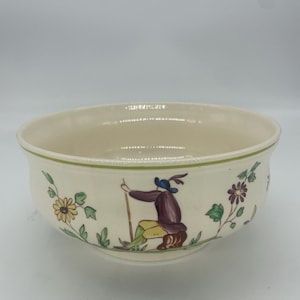 May include: Cream-colored ceramic bowl with a green rim and floral and figure illustrations. The bowl features a design of a person in a purple coat and yellow pants holding a staff. Flowers and leaves are also depicted.