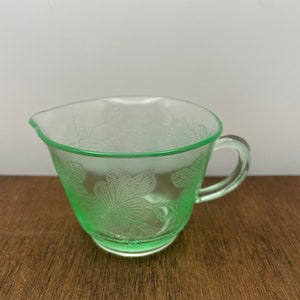 May include: A green glass creamer with a floral design. The creamer has a handle and a rounded base.
