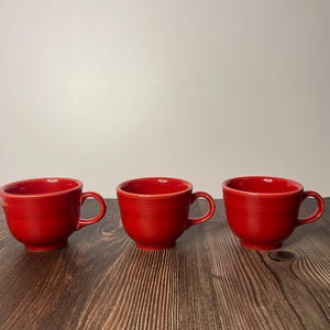 Puede incluir: Tres tazas de café de cerámica roja con asas. Las tazas están sobre una superficie de madera.