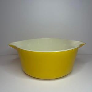 May include: A yellow ceramic mixing bowl with a white interior and rounded edges. The bowl has two handles on either side.