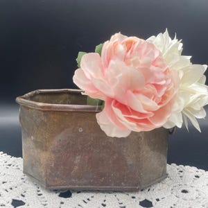 May include: A hexagonal brass planter with a distressed finish. The planter is filled with pink and white artificial flowers.