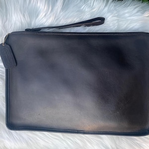 May include: A black leather clutch with a wrist strap and a zipper closure. The clutch has a rectangular shape and a small tag attached to the zipper. The background is a fluffy white surface.