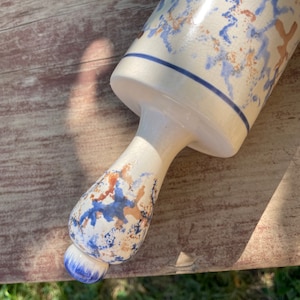 Può includere: Un mattarello in ceramica bianca con venature blu e marroni. Il mattarello ha un manico arrotondato e una fascia blu e bianca intorno alla parte superiore.