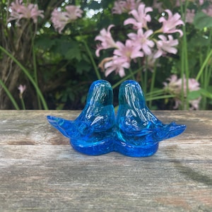 Può includere: Due statuette di uccelli in vetro blu, unite alla base, con le ali spiegate. Sono posizionate su una superficie di legno stagionato, con uno sfondo di foglie verdi e fiori rosa.