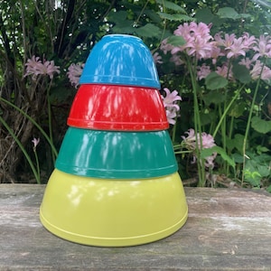 以下が含まれることがあります： 青、赤、緑、黄色の4色のミキシングボウルの積み重ね。ボウルはピラミッド型に配置され、一番大きな黄色のボウルが底にあります。ボウルは木製の表面に置かれ、背景には緑と花があります。