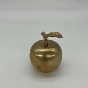 May include: A brass apple sculpture with a stem and leaf. The apple is a golden color and has a smooth, rounded shape. The stem and leaf are also brass and are attached to the top of the apple.