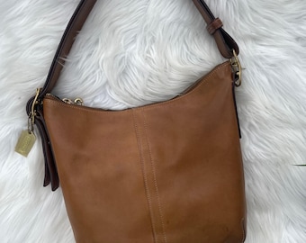 May include: A tan leather shoulder bag with a single strap and a gold-tone zip. The bag has a rectangular shape with a rounded base and a small tag attached. The bag is on a white, fluffy surface.
