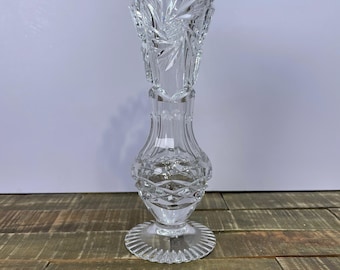 Può includere: Un vaso trasparente in cristallo tagliato con un bordo svasato e smerlato. Il vaso presenta intricati motivi a stella e a diamante, in piedi su una base rotonda e scanalata. Il vaso è alto circa 25 cm.