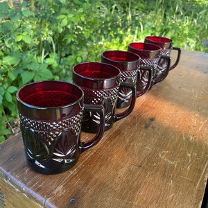 May include: Five ruby red glass mugs with a diamond pattern and a leaf design. The mugs have black handles and are lined up on a wooden surface.