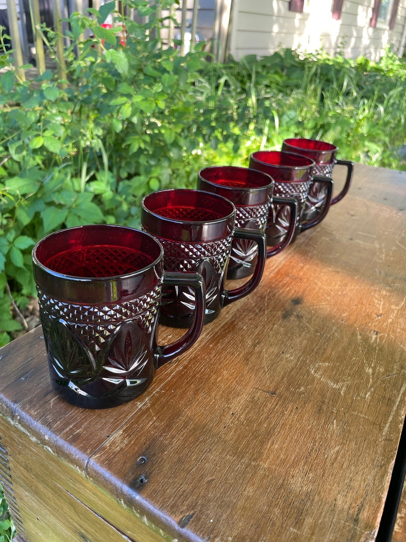 May include: Five ruby red glass mugs with a diamond pattern and a leaf design. The mugs have black handles and are lined up on a wooden surface.