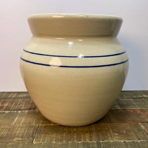 Puede incluir: Un jarrón de cerámica beige con cuerpo redondeado y borde ligeramente ensanchado. Dos rayas horizontales azul oscuro rodean el jarrón. El jarrón está sobre una superficie de madera.