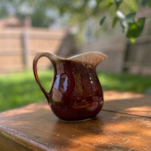 Puede incluir: Una jarra de cerámica marrón y blanca con asa. La jarra tiene un acabado esmaltado y está sentada sobre una mesa de madera.
