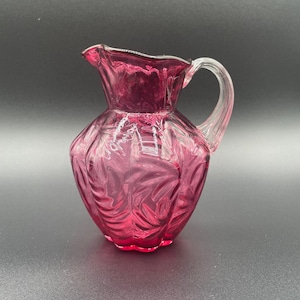 May include: A small, pink glass pitcher with a clear handle. The pitcher has a textured, floral design.