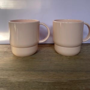 May include: Two pale pink ceramic mugs with a stacked design. The mugs have a rounded bottom and a wide, rounded handle.