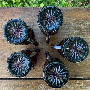 May include: Five dark red glass mugs with a starburst pattern on the bottom. The mugs are arranged in a circle on a wooden surface.