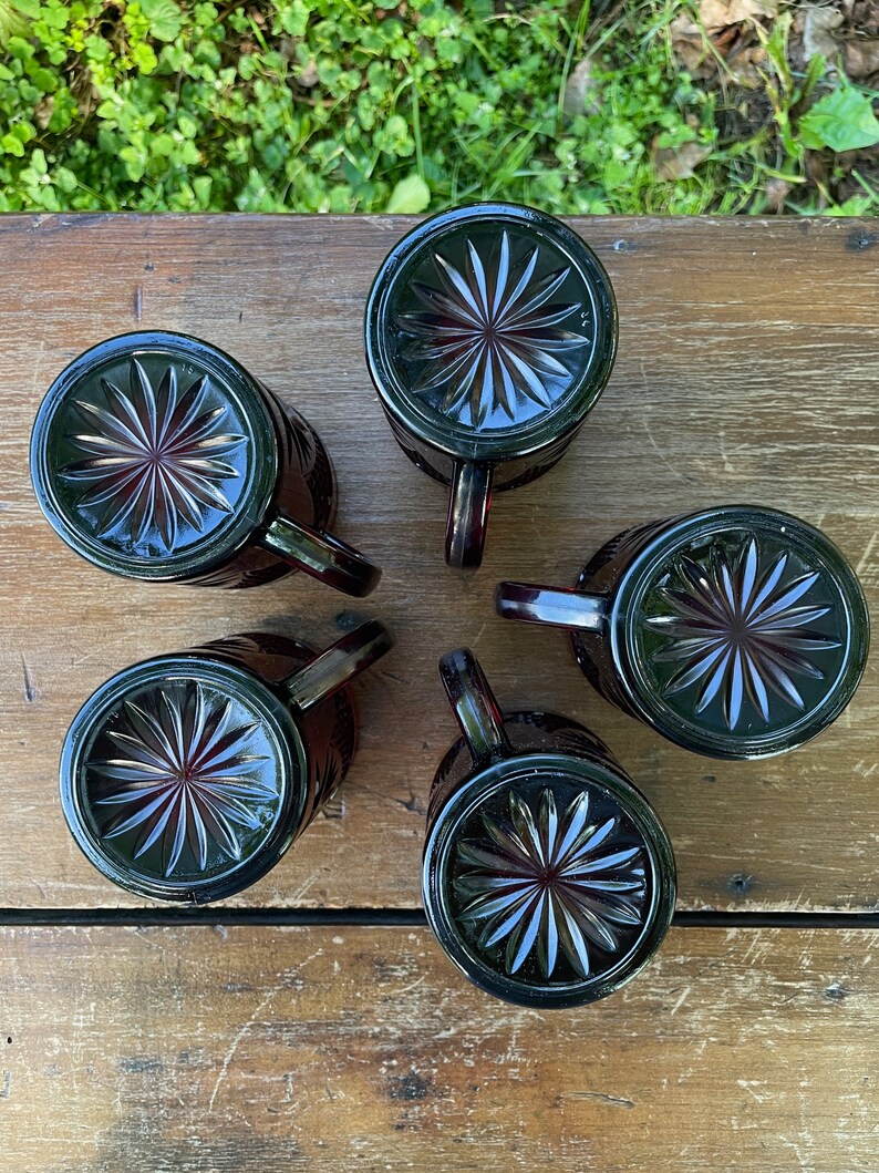 May include: Five dark red glass mugs with a starburst pattern on the bottom. The mugs are arranged in a circle on a wooden surface.