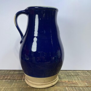 May include: A dark blue ceramic pitcher with a handle. The pitcher has a glossy finish and a light tan base. The handle is curved and attached to the side of the pitcher. The pitcher is sitting on a wooden surface.