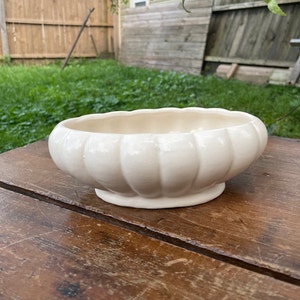 Puede incluir: Un macetero de cerámica blanco con un diseño acanalado. El macetero tiene forma ovalada y un acabado liso y brillante.