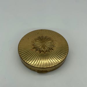May include: A vintage, round, gold-toned compact. The compact features a sunburst design on the lid with radiating lines. The compact is closed and appears to be made of metal. The overall aesthetic is classic and elegant.