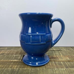 Puede incluir: Una taza de cerámica azul cobalto con base de pedestal y asa curva. La taza presenta un diseño texturizado de tejido de cesta alrededor del cuerpo. La taza está sobre una superficie de madera.