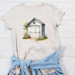 May include: A beige t-shirt with a watercolor illustration of a white shed with a door and a window. The shed is surrounded by green plants and flowers.